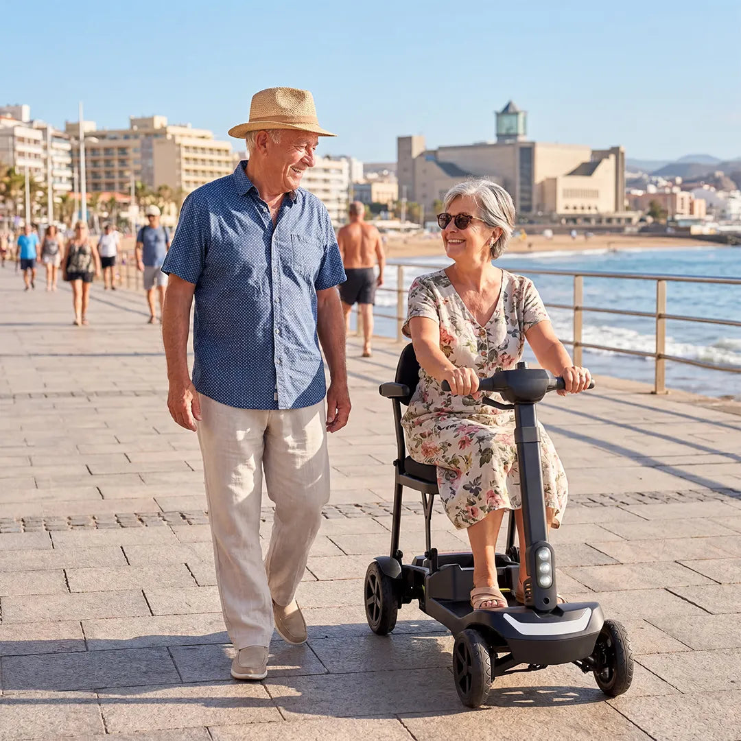 Una pareja de personas mayores sonrientes paseando por un paseo marítimo junto al mar; la mujer conduce un scooter de movilidad eléctrico de 4 ruedas ligero y de diseño moderno dortomedical