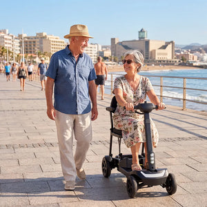 Una pareja de personas mayores sonrientes paseando por un paseo marítimo junto al mar; la mujer conduce un scooter de movilidad eléctrico de 4 ruedas ligero y de diseño moderno dortomedical